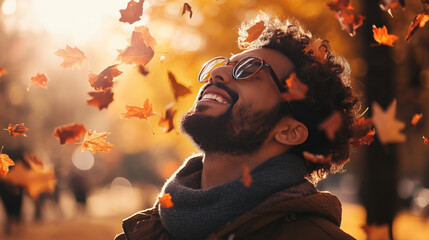 smiling ethnic man guy with eyes closed in the park outdoors in autumn surrounded by falling leaves floating in the air movement motion wearing hat curly hair glasses beard