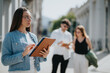 © qunica.com - Professional businesswoman using a tablet while standing outdoors, with two colleagues conversing in the background on a sunny day.
