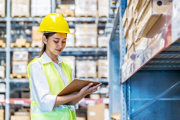 Asian woman warehouse worker in uniform doing stocktaking of products management in cardboard box on shelves in warehouse using clipboard file and pen, logistic and business export