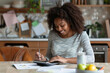 © Valeriia - Happy African-American woman calculating managing household finances at home. Focused young woman make calculations on calculator paying bills. Finance and credit concept