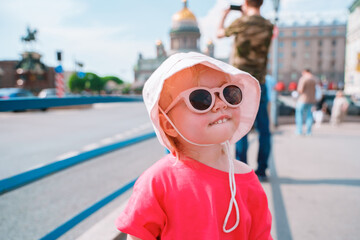 Wall Mural - A little girl in a panama hat and sunglasses walks along a city street in the center of St. Petersburg on a sunny summer day.