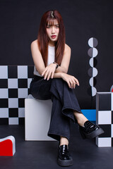 Fashion photo young confident woman model Asian beautiful female wearing trendy white sleeveless t-shirt and black jeans posing looking at camera and sitting on white box in studio colorful background