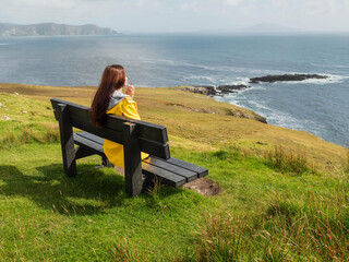 Wall Mural - A woman sits on a bench overlooking the ocean. The scene is peaceful and serene, with the woman enjoying the view. Achill island, Ireland. Popular travel and tourism area. Irish nature landscape