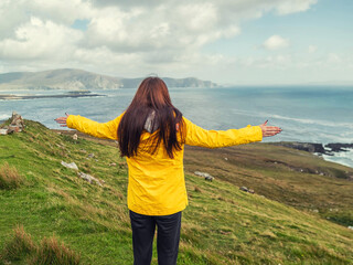 Wall Mural - Teenager girl looking at amazing Irish nature scenery with ocean and mountains of Achill island, Ireland. Travel and tourism. Popular travel and tourism area with stunning view. Warm day, cloudy sky.