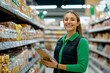 © SejutaCahaya - A smiling female supermarket employee stands in an aisle with a clipboard in hand, looking at the camera.