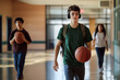 © SejutaCahaya - A teenage boy walks down a high school hallway with a basketball and headphones on, two other students in the background.