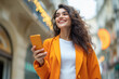 © Niks Ads - young beautiful woman wearing orange jacket smiling and holding smartphone while walking