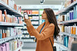 © SejutaCahaya - A young woman uses a smartphone to compare prices while shopping for hair care products in a supermarket aisle.