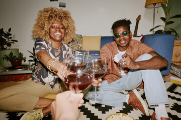 Biracial company of friends drinking red wine and giving toast together while sitting on black white carpet