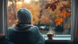 © ISVO - woman in a knitted hat and sweater looking out of a window with a cup of coffee on the windowsill and autumn leaves in the rain