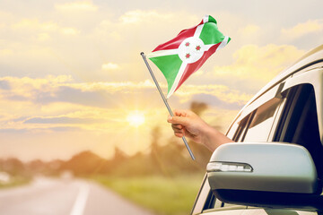 Waving the Burundi flag against the sunrise or sunset from a car driving along a country road. Holding the Burundi flag, traveling by car, on a weekend trip.