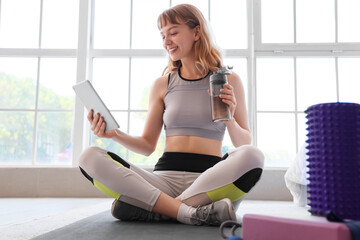 Poster - Young sporty woman with tablet and bottle of water resting after training at home