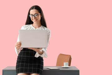 Pretty young female programmer with laptop sitting on table against pink background