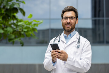 Wall Mural - Smiling doctor with stethoscope and glasses using smartphone outside hospital, highlighting modern healthcare, technology, communication, and professionalism.