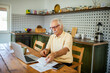 © Marko Geber - Elderly man managing bills and finances on laptop in kitchen