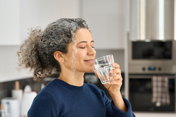 Happy healthy woman of middle age holding glass of water standing at home. Mature 50 years old lady enjoying drinking pure mineral clear water for body hydration. Morning daily hydrating treatment.