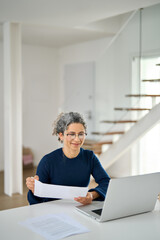 Mature lady paying bills online checking financial papers invoice making baking payments using computer. Middle aged older woman looking at laptop holding documents hybrid working at home table.