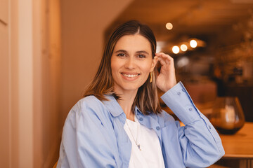 Canvas Print - Cute brunette short hair girl smiling and fix hair. Woman fix her hair, look at camera. She wear blue shirt, white top, sitting rest in cafe. Happy brunette girl look flirting and coquettish.