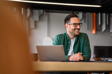 Wall Mural - happy young businessman in eyeglasses looking away while working online over laptop at desk in offic