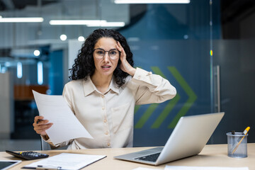 Wall Mural - Hispanic businesswoman looking stressed while sitting at office desk holding report. Laptop open in modern workspace, expressing concern. Professional setting suggests business environment with