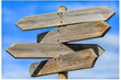 © Avelino - A wooden signpost with four blank arrows pointing in different directions, against a blue sky with clouds, representing a choice or a crossroads. Generated with AI.