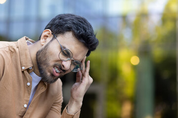 Young man with glasses looks frustrated, experiencing headache or stress. Outdoor setting captures emotion and discomfort. Hand on head indicates pain or tension.