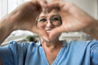 © fizkes - Mature woman wears eyeglasses looking at camera through joined fingers showing heart symbol, close up. Advertises eyewear store, laser vision correction, health check-up, cardio vitamins, sign of love