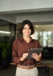 © insta_photos - Smiling young Hispanic professional business woman using tablet standing in office. Happy businesswoman manager, female executive leader holding tab at work, looking at camera. Vertical portrait.