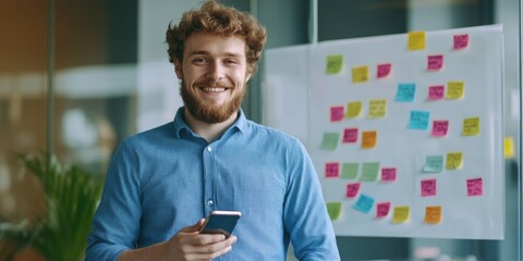 Wall Mural - A man with a beard is smiling and holding a cell phone. He is standing in front of a white board with colorful sticky notes on it