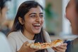 © vefimov - A woman is smiling and holding a slice of pizza. She is surrounded by other people, and they are all laughing and enjoying their time together. The atmosphere is lighthearted and fun