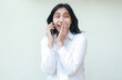 © M Alfan Setyawan - image of delighted asian young woman secretary speaking on smartphone looking away covering mouth with palms feeling shy talking gossip whispering wearing white formal suit shirt standing isolated