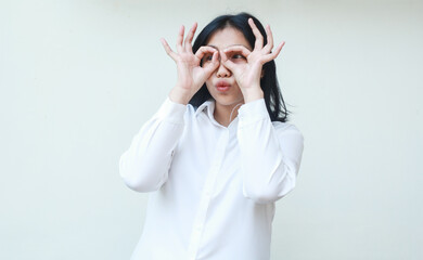 happy satisfied young asian woman showing ok signs on eyes wearing white formal suit shirt feeling proud looking away, okay symbols, standing over isolated background