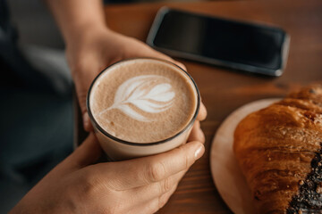 A woman holding a coffee cup with a leaf design on it. She is sitting at a table with a croissant and a cell phone.