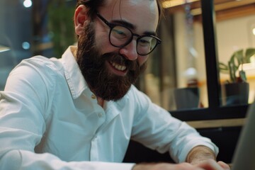 Wall Mural - A man with a beard and glasses is smiling while using a laptop. He is enjoying himself and is likely working or browsing the internet