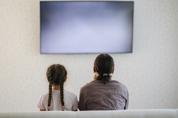 Back view of teenage boy with his six year old sister watching TV in living room. Big screen 