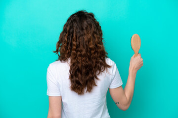 Young caucasian woman holding hairbrush isolated on blue background in back position