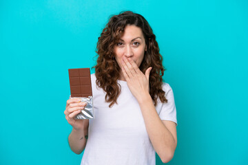 Wall Mural - Young caucasian woman isolated on blue background taking a chocolate tablet and surprised