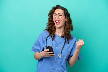 Wall Mural - Young nurse caucasian woman isolated on blue background with phone in victory position