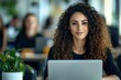 © Fathor - Beautiful Middle Eastern Manager Sitting at a Desk in Creative Office. Young Stylish Female with Curly Hair Using Laptop Computer in Marketing Agency. Colleagues Working in the Background.generative a