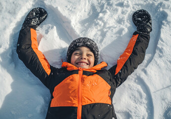 Canvas Print - A young boy in an orange and black jacket, lying on the snow with his arms outstretched to form a heart shape while making a snow angel