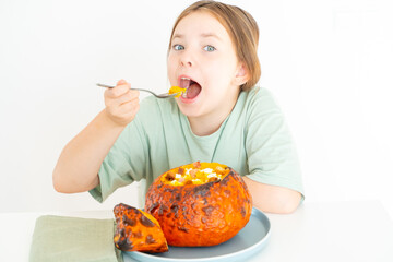 Cute, little teenage girl eats pumpkin soup served in baked pumpkin. Happy Girl in a green T-shirt with an autumn dish for Halloween on a white background.