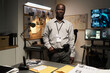 © pressmaster - Portrait of African American FBI agent wearing badge, standing in a dimly lit office with various screens and bulletin board in background examining documents