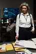 © pressmaster - Gray-haired woman with FBI badge reviewing documents at office desk, team member analyzing data on monitors in background