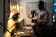 © pressmaster - Black man pouring coffee into cup in dimly lit office filled with various work-related equipment and documents hanging on wall. Cozy and focused workspace setting