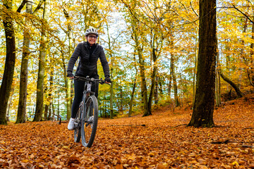 Wall Mural - Middle aged woman wearing black sporty jacket, black cycling pants and white bike helmet riding bicycle in forest in autumn scenery. Front view.