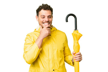 Wall Mural - Brazilian man with rainproof coat and umbrella over isolated chroma key background looking to the side and smiling