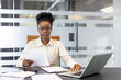 © Liubomir - Professional African American businesswoman engaged in work at office desk. Concentrated on reviewing documents with laptop open. Represents corporate environment, focus, and business tasks.