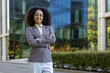 © Liubomir - Confident African American businesswoman standing with arms crossed outside office building. She wears formal business attire symbolizing empowerment, leadership, and professionalism.