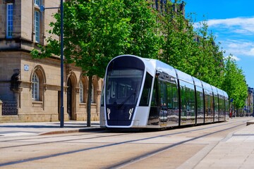 Luxembourg, A modern silver tram moves swiftly along the street, creating a motion blur, while a vintage stone building with many windows stands alongside, surrounded by trees and urban scenery.