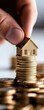 © Rossarin - A hand placing a miniature house on a stack of gold coins, symbolizing investment, wealth, and real estate market concepts.